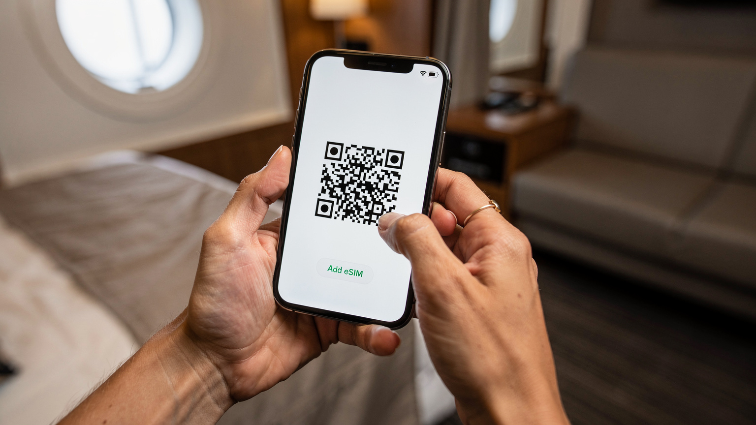 A woman's hands, with a subtle wedding band, holding a modern smartphone, displaying an eSIM activation QR code on the screen, against a blurred background of a ship's cabin or lounge area.
