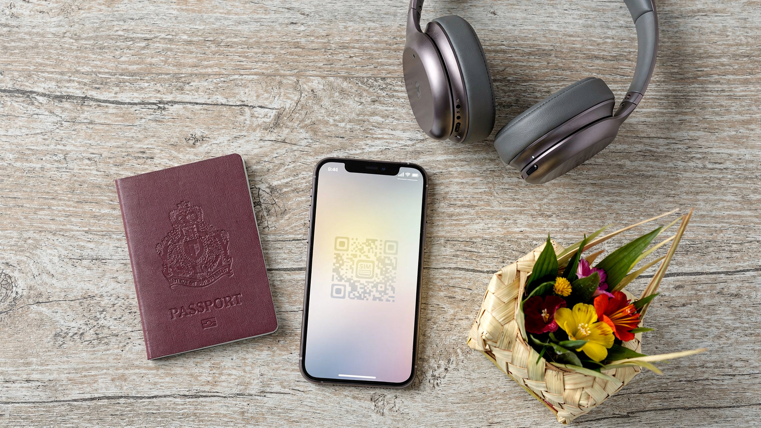An editorial overhead flat-lay shot featuring a passport, a smartphone with an eSIM QR code partially visible, a pair of headphones, and a small, traditional Balinese woven basket with offerings, all on a light wooden table.