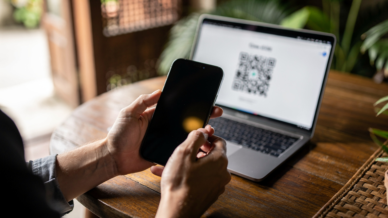 Close-up of hands holding a smartphone, a QR code for eSIM activation displayed on a laptop screen in the background, set in a cozy Balinese-style cafe.