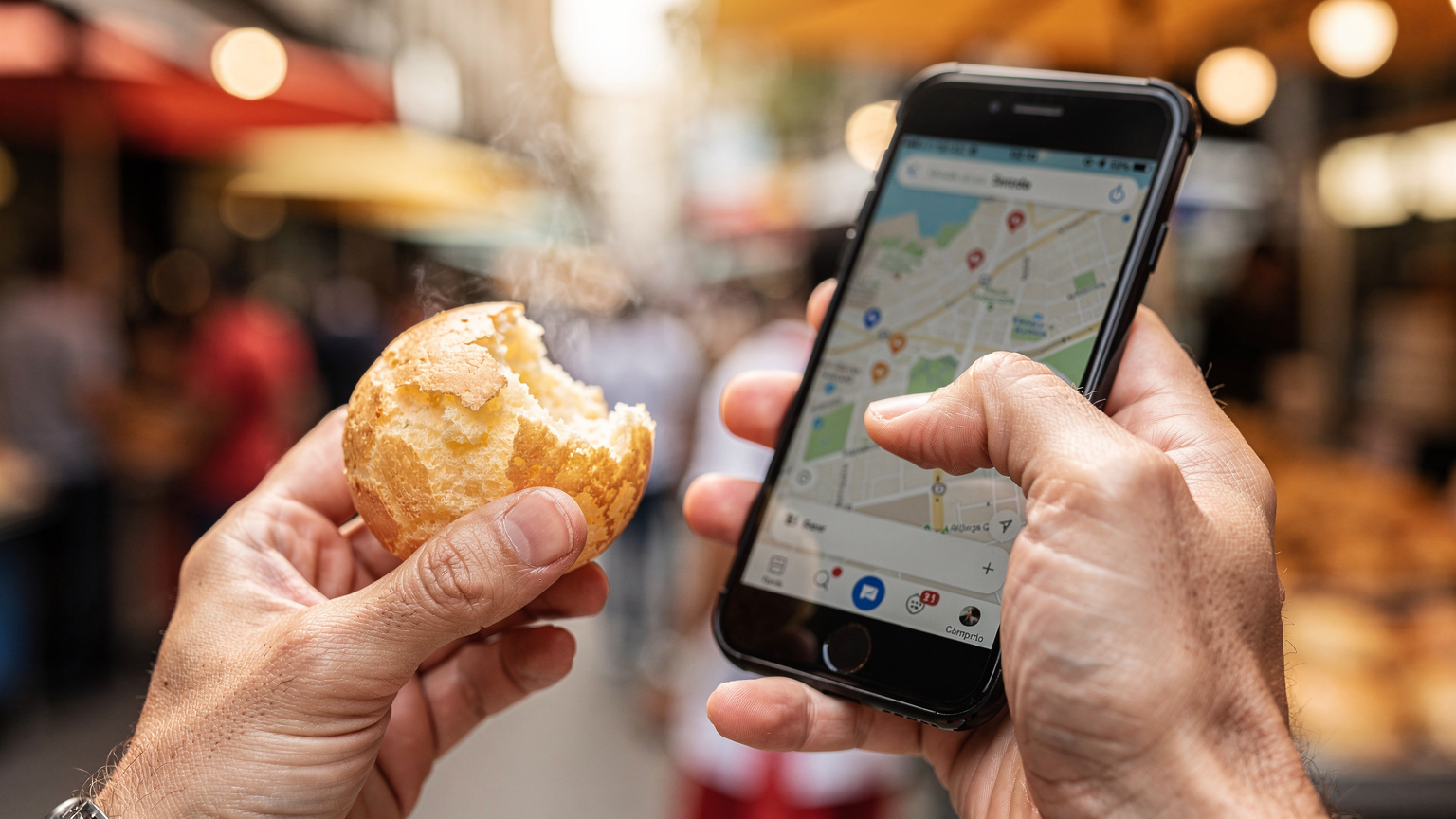 Close-up of a traveler's hands interacting with their smartphone screen, displaying a map application, while holding a traditional Brazilian Pão de Queijo in the other hand.