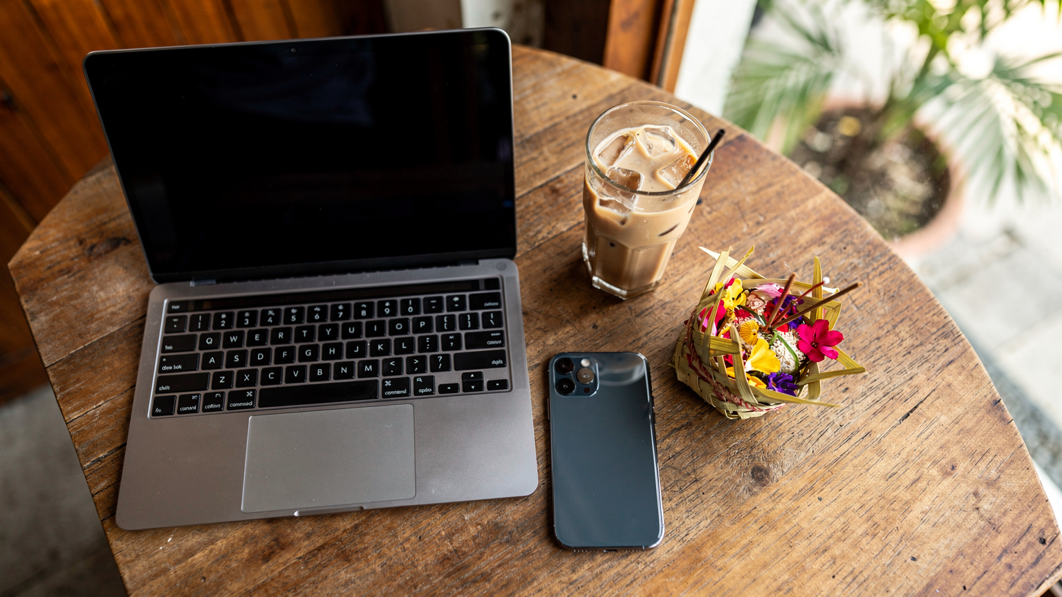 Uma mesa de café em Canggu, Bali, com um laptop aberto, um iPhone 17, um copo de café gelado e uma pequena oferenda Balinesa (Canang Sari), mostrando um ambiente de trabalho descontraído.