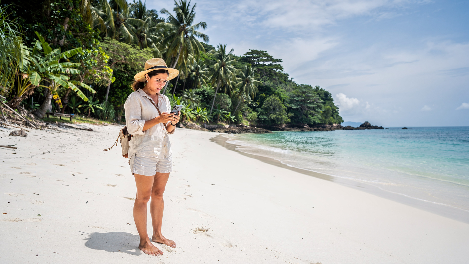 A solo female traveler standing on a beautiful, secluded beach in Thailand, looking at her smartphone with a frustrated expression as the screen shows a 'no signal' icon, with lush tropical jungle in the background.