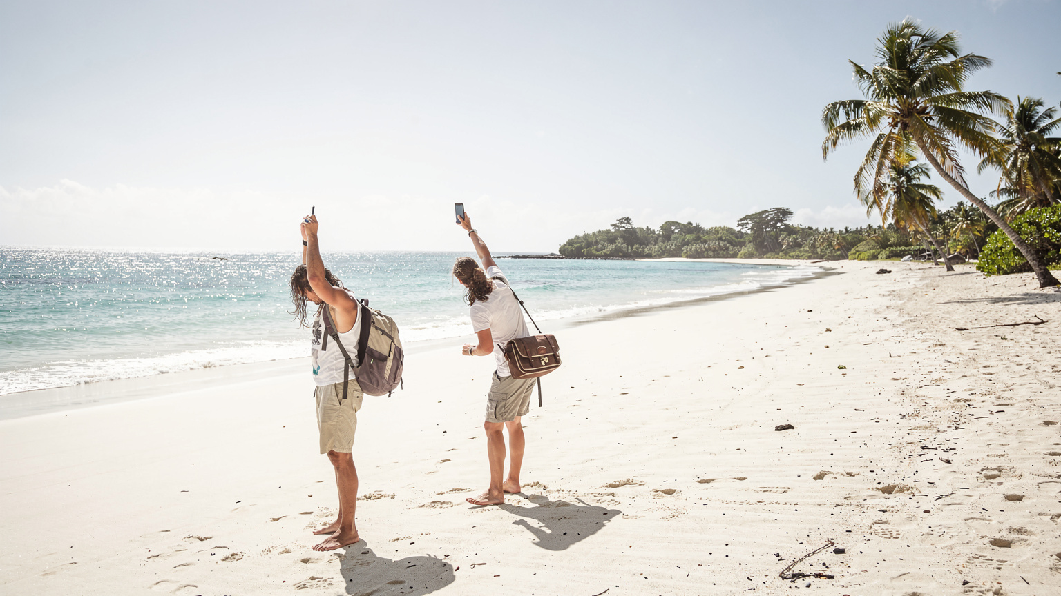 Seorang traveler dengan ekspresi cemas memegang ponselnya tinggi-tinggi, mencoba mencari sinyal di tepi pantai terpencil yang sepi di Fiji.