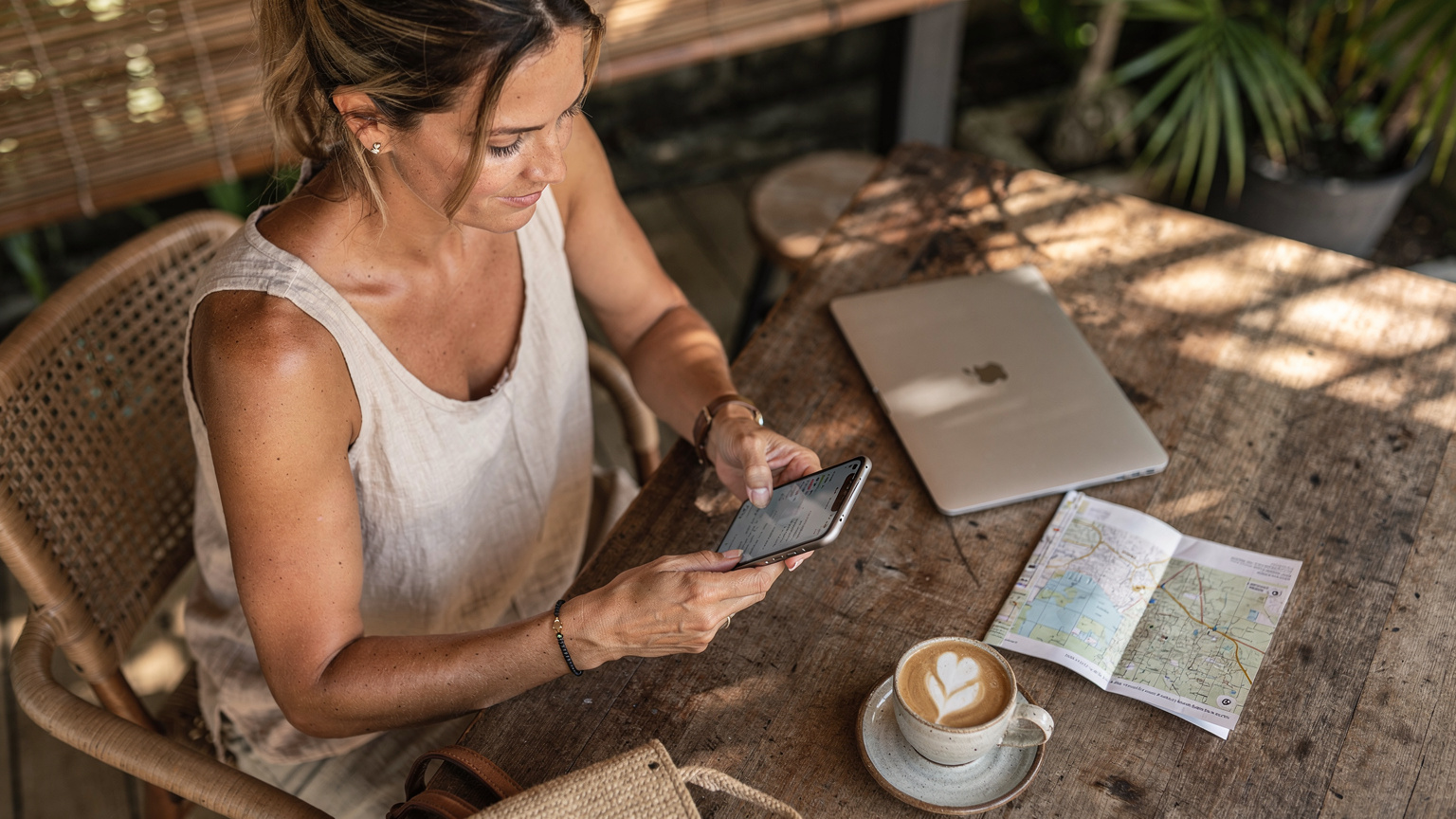 Seorang backpacker sedang membandingkan harga paket eSIM di iPhone 17 miliknya yang terbuka di kafe hipster di Canggu, Bali, dikelilingi oleh laptop dan cangkir kopi.