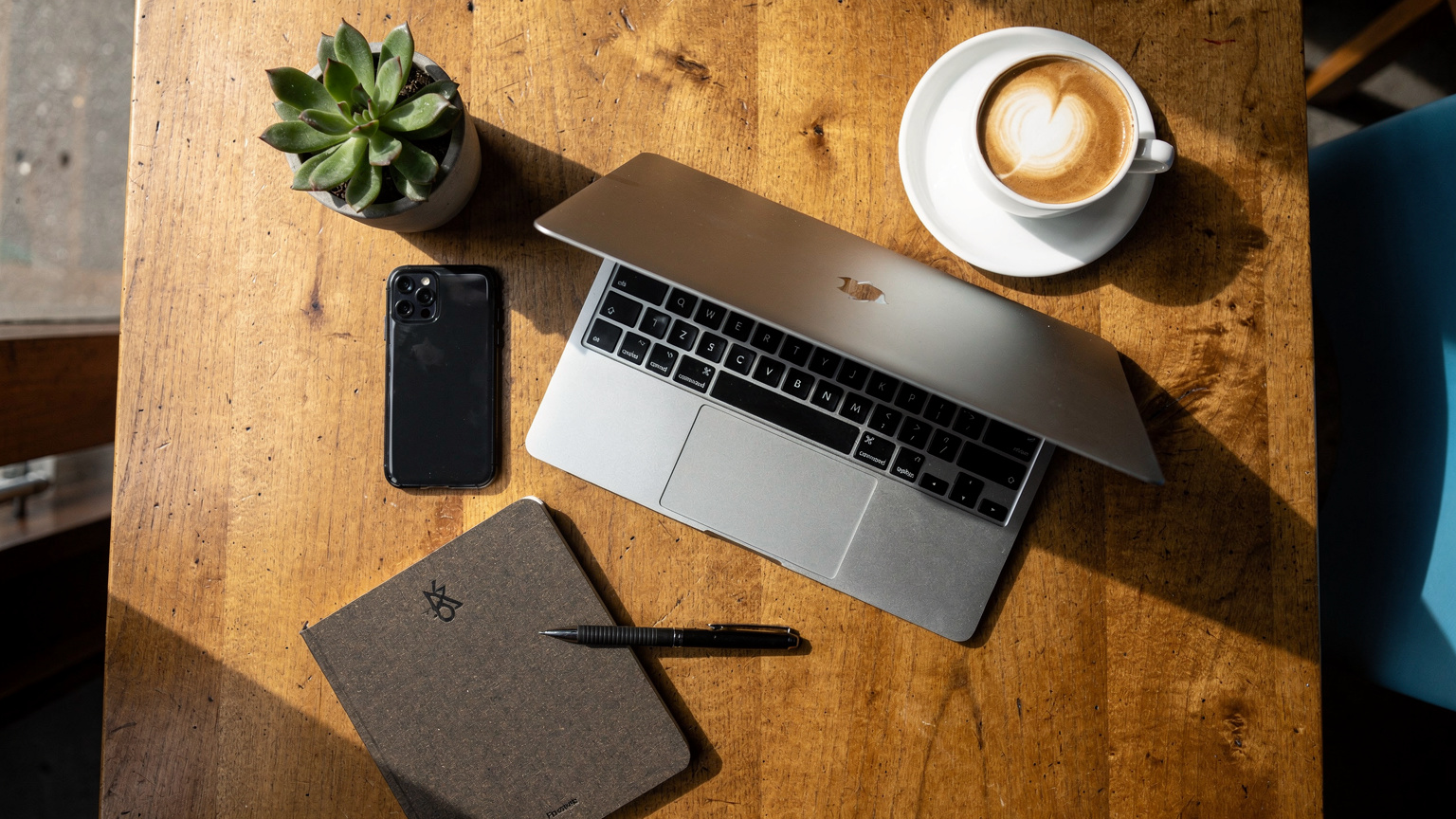 An overhead flat-lay of a digital nomad's workspace in a stylish Cape Town cafe, showing a laptop, smartphone, coffee cup, and a small, locally-branded notebook.