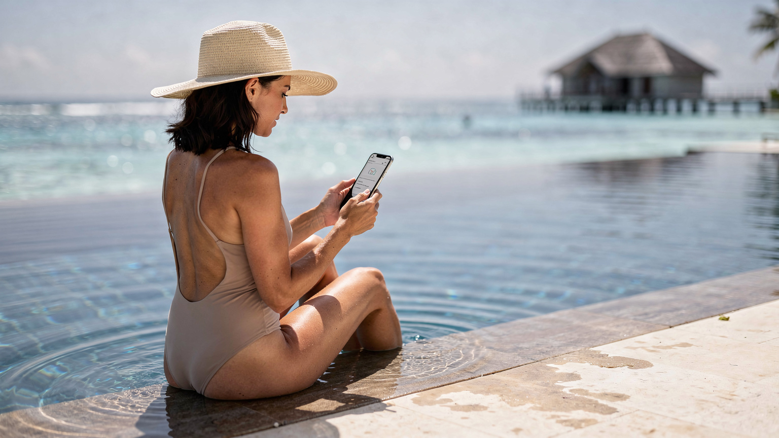 Una donna seduta a bordo piscina in un resort maldiviano, con il telefono in mano che mostra un'applicazione di monitoraggio del consumo dati, sullo sfondo le acque turchesi.