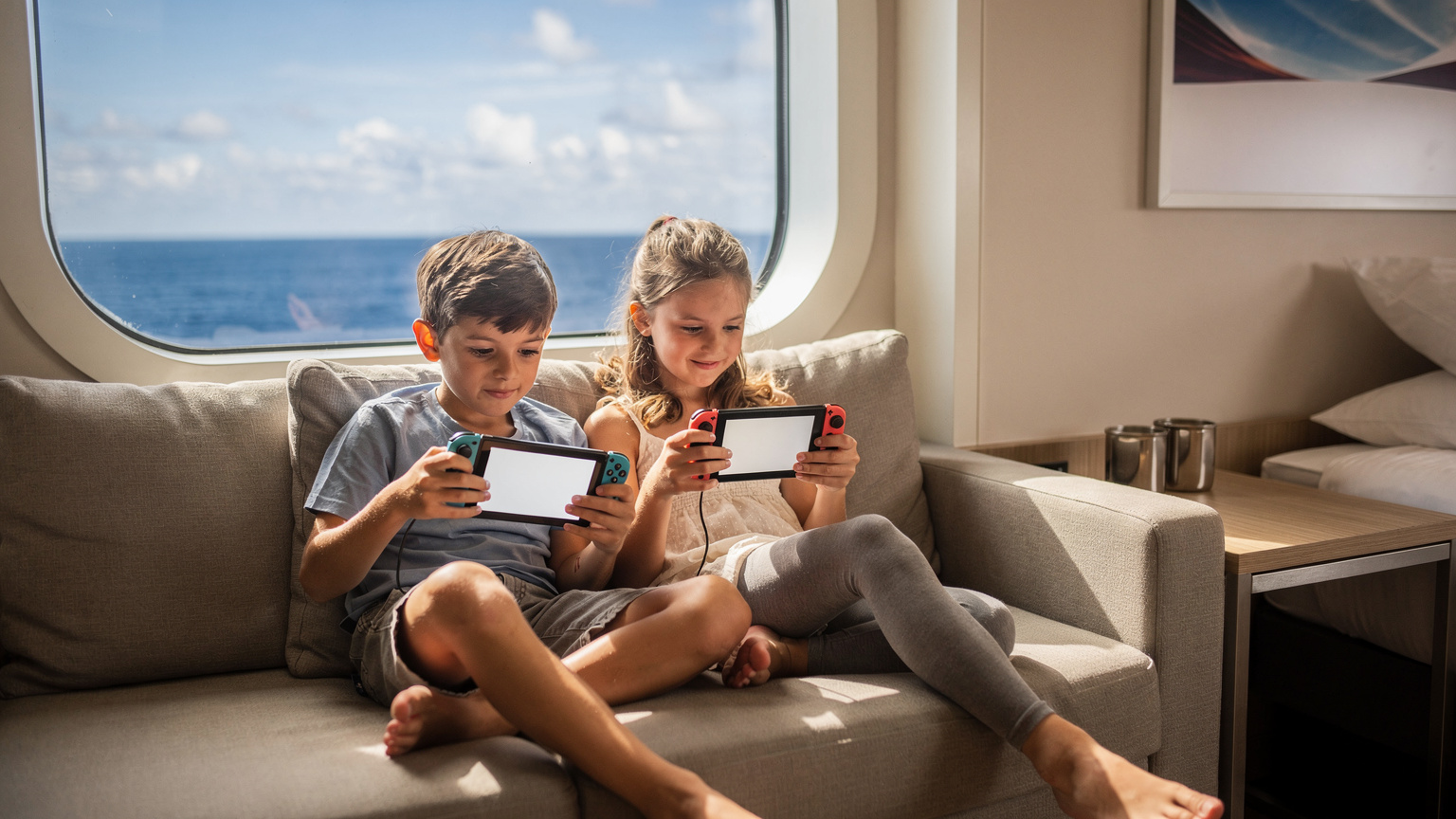 Seorang anak laki-laki dan perempuan asyik bermain game di Nintendo Switch mereka di dalam kabin kapal pesiar, dengan pemandangan laut di luar jendela, menunjukkan konektivitas bahkan di tengah laut.