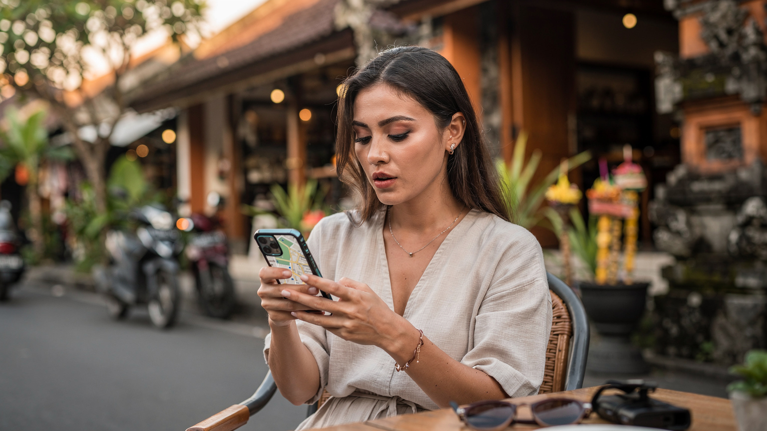 Seorang wanita muda duduk di kafe trendi di Seminyak, sedang melihat peta digital di ponselnya, dengan latar belakang jalanan yang ramai dan butik-butik yang menarik.